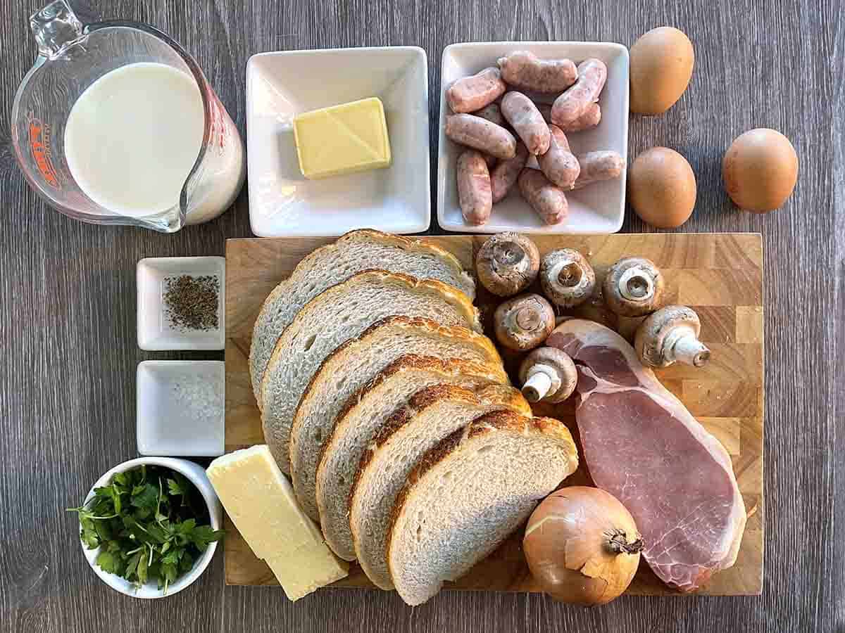 ingredients on a board including bread, sausage, bacon, onion, cheese, egg,s milk butter and parsley.